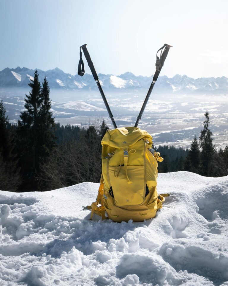 A bright yellow backpack and hiking poles stand on a snowy mountain landscape, showcasing winter adventure.
